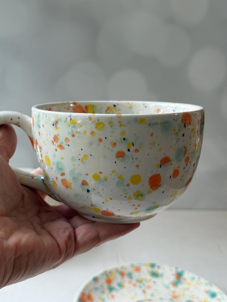 May include: A white ceramic teacup and saucer set with a speckled design in orange, yellow, blue, and green. The teacup has a large handle and a wide bowl. The saucer is visible at the bottom.