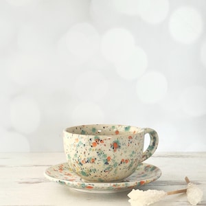 May include: A ceramic teacup and saucer set with a speckled design. The cup and saucer are off-white with colorful splatters of orange, blue, and green. The cup has a handle and sits on a matching saucer. The set is on a white wooden surface.