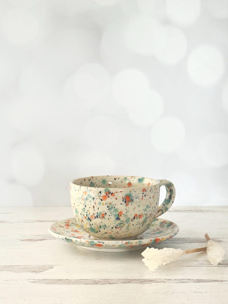 May include: A ceramic teacup and saucer set with a speckled design. The cup and saucer are off-white with colorful splatters of orange, blue, and green. The cup has a handle and sits on a matching saucer. The set is on a white wooden surface.