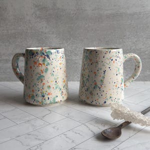 May include: Two ceramic mugs with a speckled design in shades of blue, green, and orange on a cream background. Each mug has a curved handle and a gold rim. The mugs are displayed on a white marble-patterned surface.