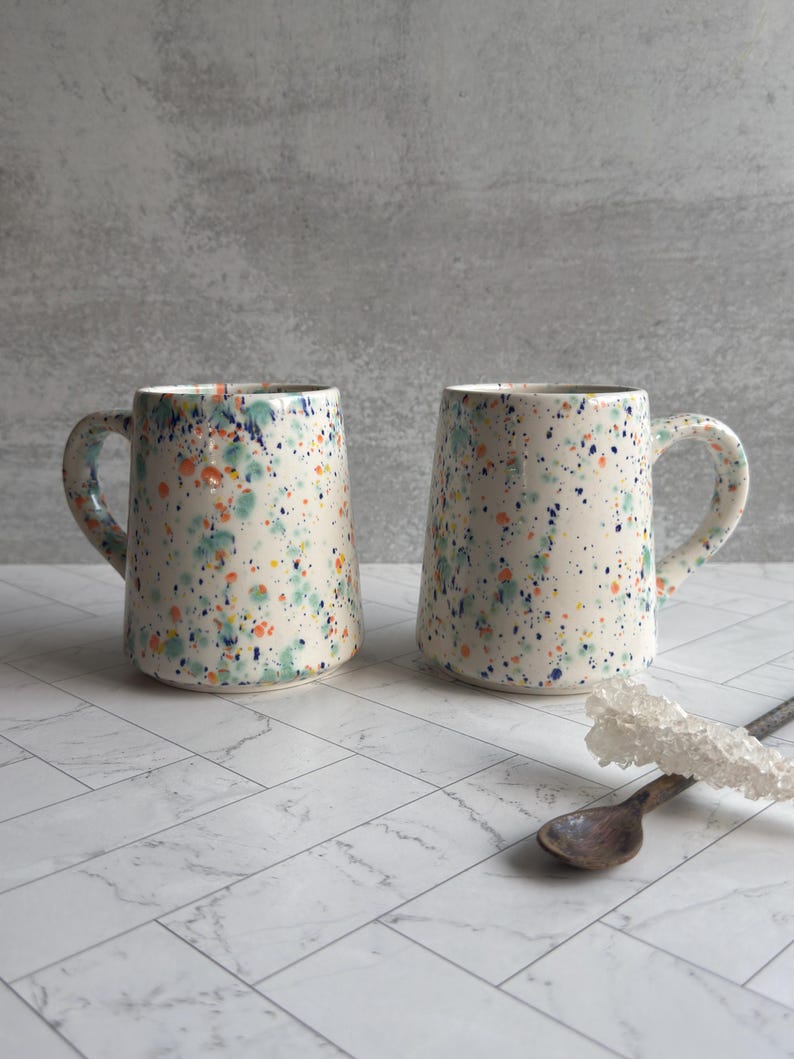 May include: Two ceramic mugs with a speckled design in shades of blue, green, and orange on a cream background. Each mug has a curved handle and a gold rim. The mugs are displayed on a white marble-patterned surface.