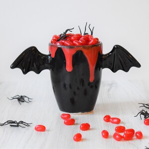 May include: Black ceramic mug with bat wings, filled with red jelly beans and a red glaze dripping down the sides.