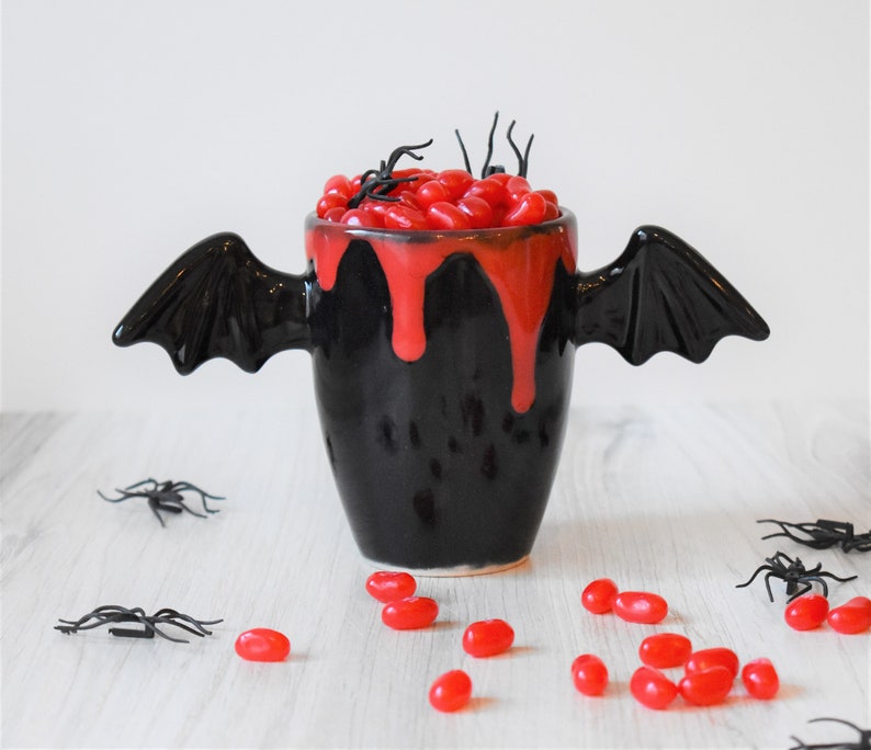 May include: Black ceramic mug with bat wings, filled with red jelly beans and a red glaze dripping down the sides.