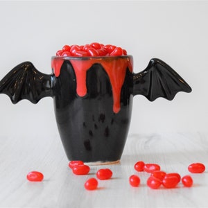 May include: A black ceramic mug with bat wings, filled with red jelly beans and dripping red glaze. The mug is sitting on a white wooden surface.