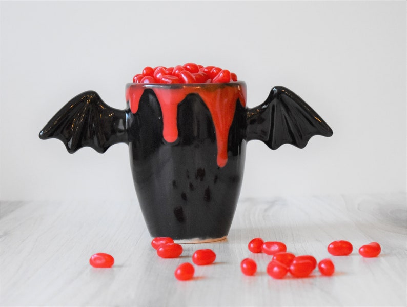 May include: A black ceramic mug with bat wings, filled with red jelly beans and dripping red glaze. The mug is sitting on a white wooden surface.