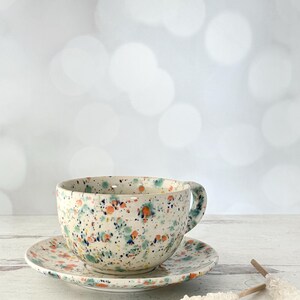May include: A speckled ceramic teacup and saucer set. The cup and saucer are white with a colorful splatter pattern of orange, blue, and green. The cup has a handle and sits on a matching saucer.