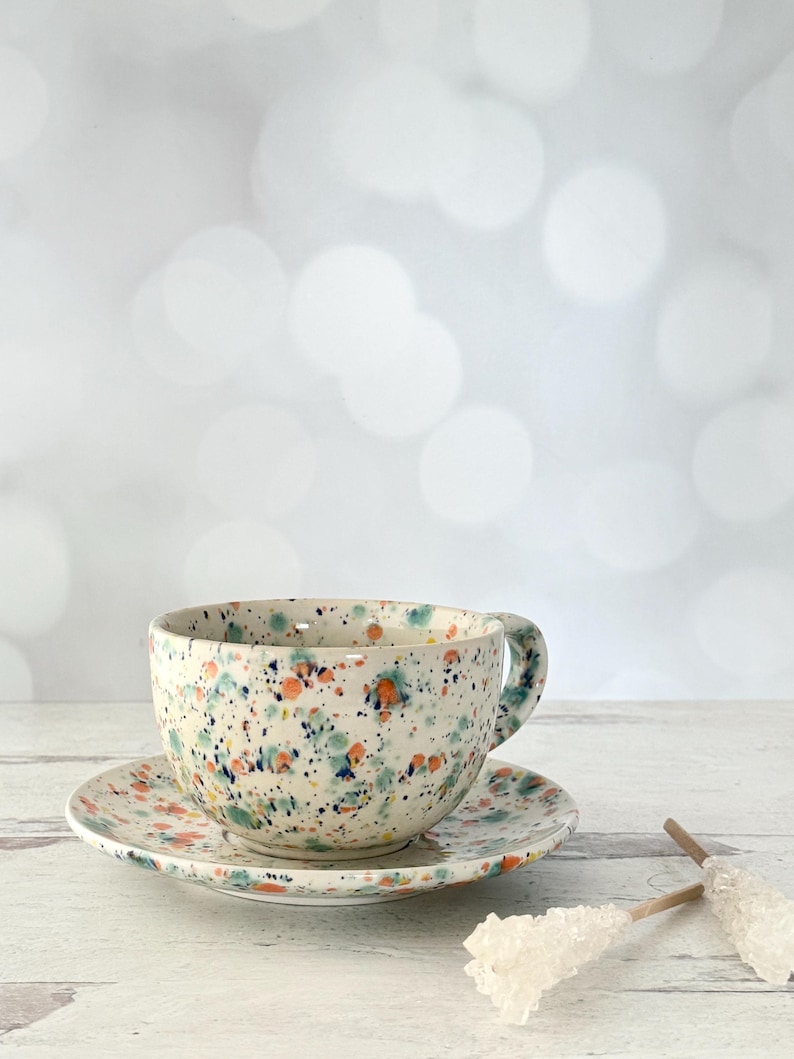 May include: A speckled ceramic teacup and saucer set. The cup and saucer are white with a colorful splatter pattern of orange, blue, and green. The cup has a handle and sits on a matching saucer.