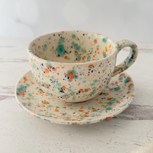 May include: A ceramic teacup and saucer set. The off-white cup and saucer are decorated with colorful paint splatters in shades of blue, orange, and green. The cup has a curved handle.