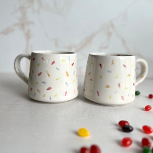 May include: Two white ceramic mugs with a confetti pattern in shades of red, yellow, blue, and green. The mugs have a rounded shape and a handle on one side.