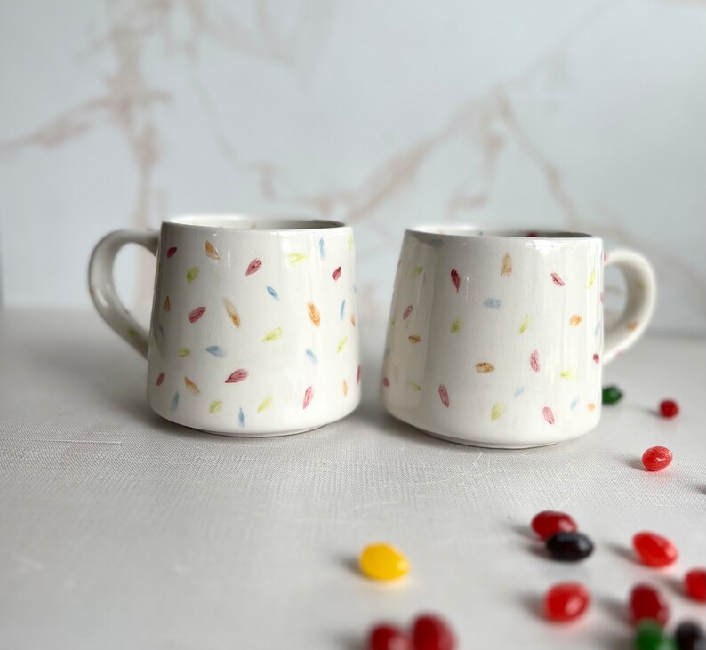 May include: Two white ceramic mugs with a confetti pattern in shades of red, yellow, blue, and green. The mugs have a rounded shape and a handle on one side.