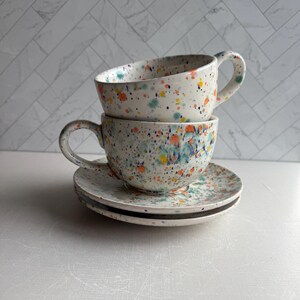 May include: Two ceramic teacups and saucers stacked. The cups and saucers are white with colorful paint splatters in blue, orange, yellow, and green. The cups have large handles.