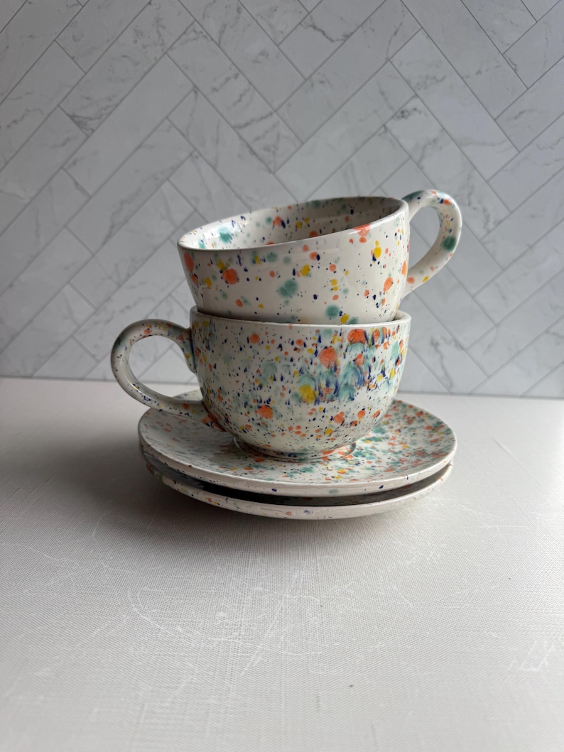 May include: Two ceramic teacups and saucers stacked. The cups and saucers are white with colorful paint splatters in blue, orange, yellow, and green. The cups have large handles.