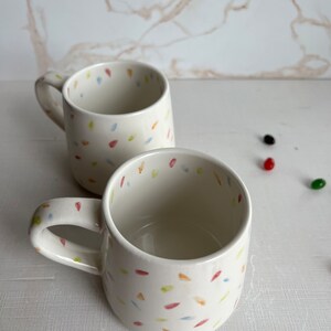 May include: Two white ceramic mugs with a confetti pattern in shades of blue, yellow, orange, and red. The mugs have a rounded shape and a handle.