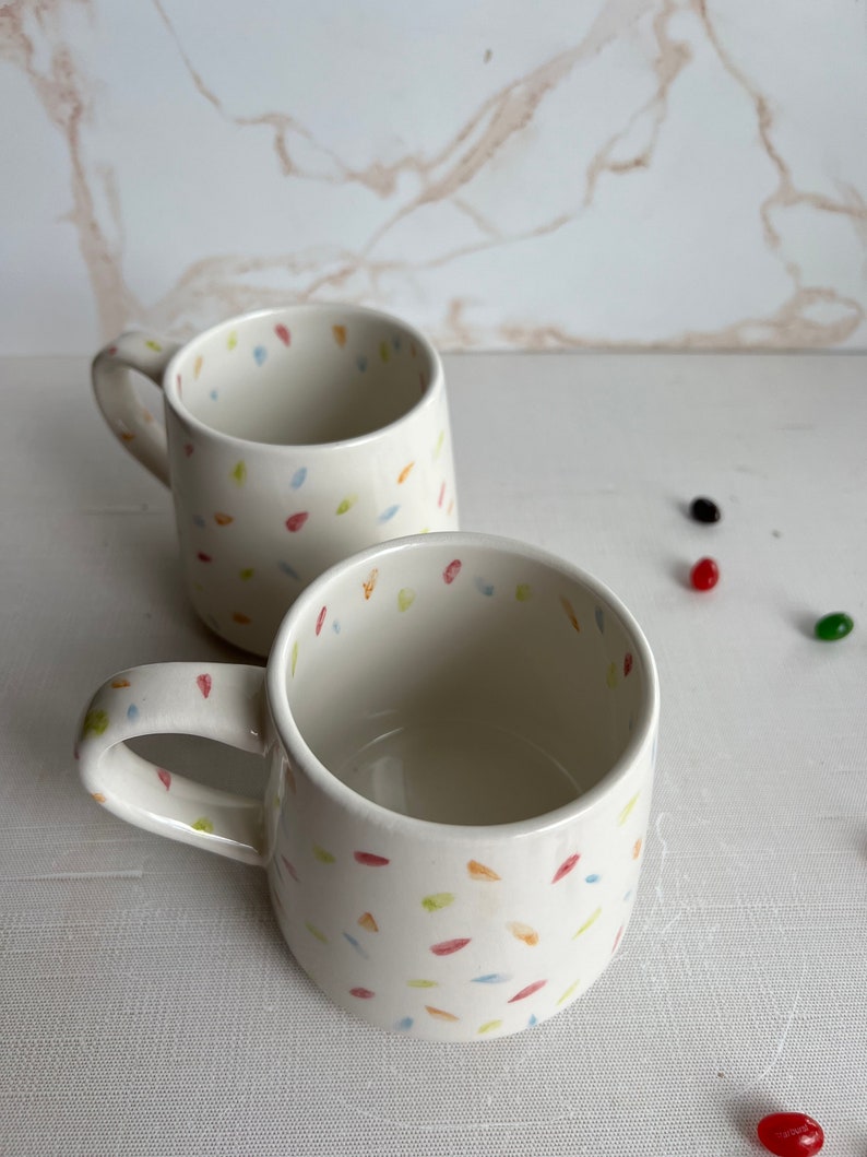 May include: Two white ceramic mugs with a confetti pattern in shades of blue, yellow, orange, and red. The mugs have a rounded shape and a handle.