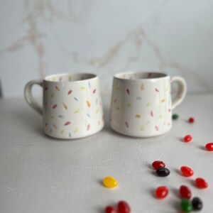 May include: Two white ceramic mugs with a confetti pattern in shades of pink, yellow, blue, and green. The mugs have a handle on the right side. The mugs are sitting on a white surface with scattered jelly beans in various colors.