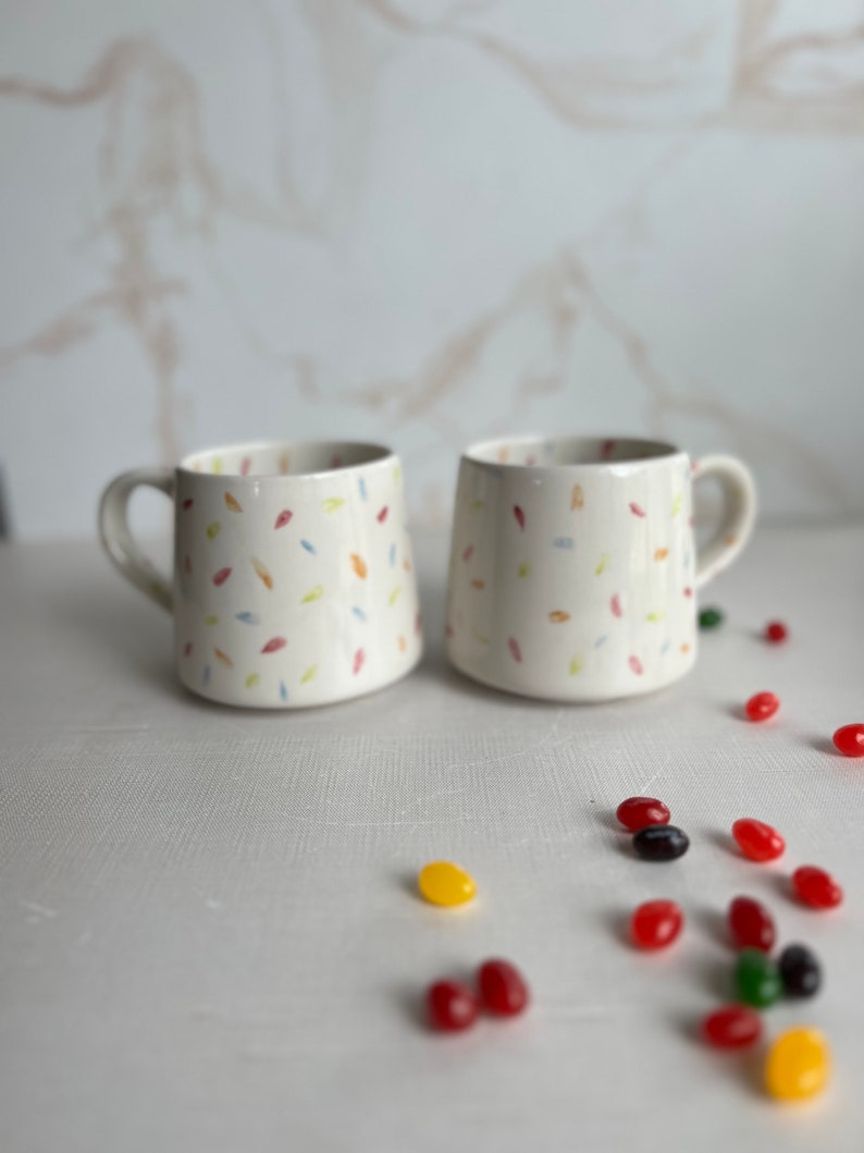 May include: Two white ceramic mugs with a confetti pattern in shades of pink, yellow, blue, and green. The mugs have a handle on the right side. The mugs are sitting on a white surface with scattered jelly beans in various colors.