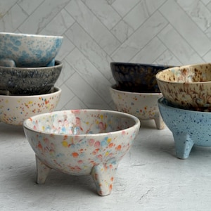 May include: A collection of ceramic bowls with speckled patterns and three small legs. The bowls come in various colors, including blue, white, and brown. The speckled designs feature colorful dots.