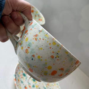 May include: White ceramic teacup and saucer set with a speckled design in orange, yellow, and blue. The teacup has a curved handle and is partially filled. The saucer is visible below the cup.