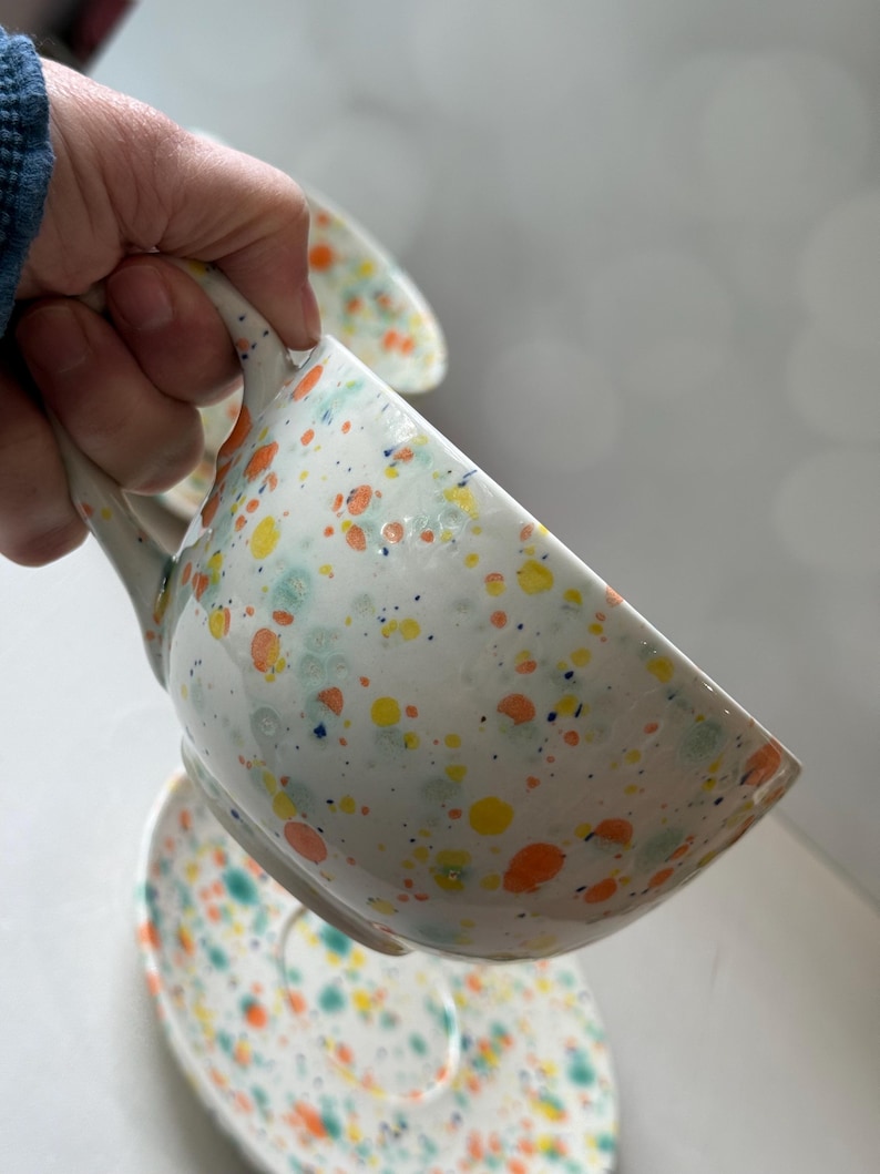 May include: White ceramic teacup and saucer set with a speckled design in orange, yellow, and blue. The teacup has a curved handle and is partially filled. The saucer is visible below the cup.