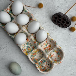 May include: A ceramic egg holder with a speckled design in orange, yellow, and blue holds seven white eggs. One speckled blue-green egg rests beside the holder. Dried floral elements are in the background. The egg holder is a kitchenware item.