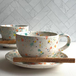 set of two white ceramic teacup and saucer with a speckled pattern of yellow, orange, and green. latte mug