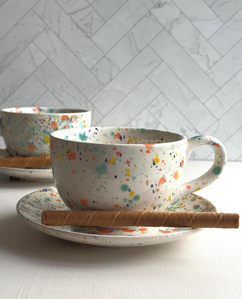 set of two white ceramic teacup and saucer with a speckled pattern of yellow, orange, and green. latte mug
