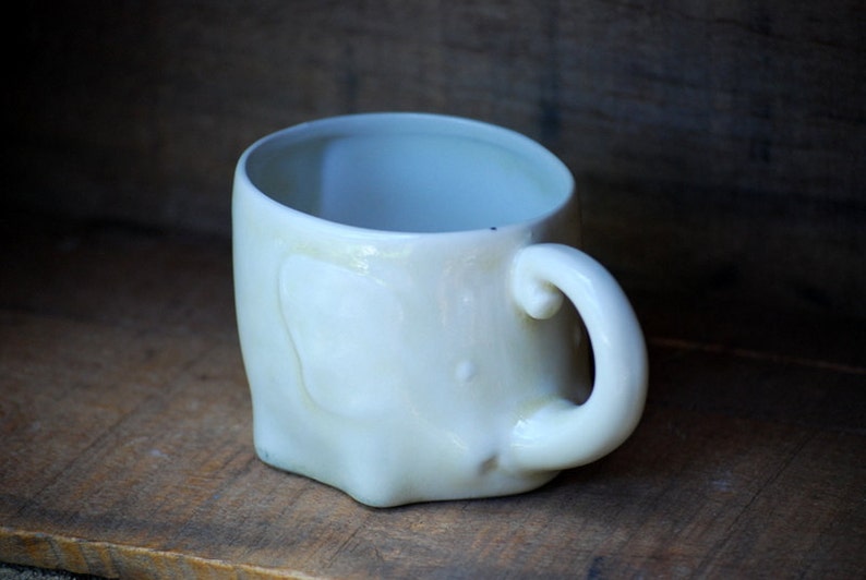 Puede incluir: Una taza de cer&aacute;mica blanca rota hecha a mano, con una forma redondeada &uacute;nica. La taza presenta un asa curvada y una superficie ligeramente irregular, lo que le da un aspecto r&uacute;stico y artesanal. La taza mide aproximadamente 10 cm de alto.