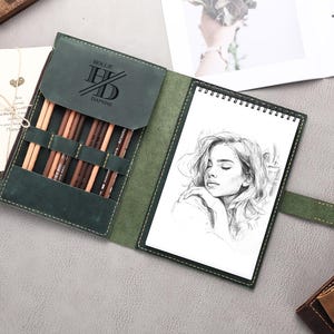 May include: A green leather sketchbook with a zipper closure and a built-in pencil holder. The sketchbook has a personalized inscription that reads "Hollie & Daphne".