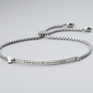 Puede incluir: Una pulsera plateada con una cadena fina y un cierre deslizante. La pulsera presenta una barra rectangular grabada con la frase "I love you to the moon and back". Un dije en forma de corazón está adjunto.