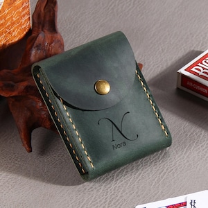 May include: A dark green leather wallet featuring a flap closure with a brass-colored metal snap button. The front of the wallet is embossed with the letter N and the name Nora. It is constructed with visible thick tan stitching along the edges.