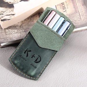 May include: A green leather pen case with a flap closure. The case has the initials "K+D" and the names "Kendra" and "Denise" engraved on the front. The case is filled with five pens.