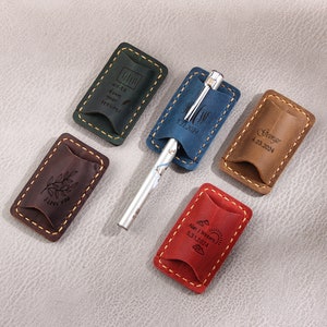May include: Five leather pen holders with different engraved designs. The holders are brown, green, blue, red, and tan. The designs include a branch with leaves, a mountain range, a pen and the date 5.31.2024, a pen and the date 4.23.2024, and a pen and the date 5.31.2024.