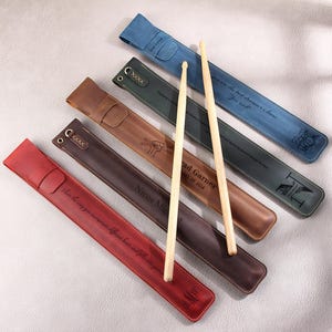 May include: Six leather-wrapped drumsticks with personalized engravings. The drumsticks are brown, red, blue, and black. The engravings include names, dates, and messages.