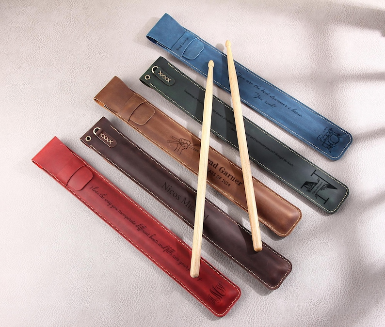 May include: Six leather-wrapped drumsticks with personalized engravings. The drumsticks are brown, red, blue, and black. The engravings include names, dates, and messages.