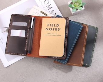 May include: Four leather notebook covers in different colors: brown, dark blue, light blue, and dark brown. The covers have a stitched edge and are designed to hold a Field Notes notebook. The notebook cover on the left has two card slots and a pen holder.