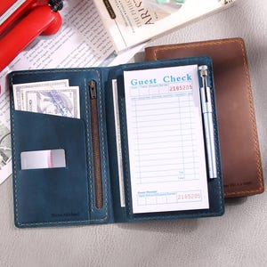 May include: A blue leather wallet with a zipped pocket and a guest check notepad. The notepad has a brown leather cover and the text "Guest Check" printed on it. The wallet has the text "Nixon Michael" printed on the front.