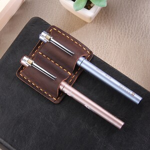 May include: A brown leather pen holder with two slots for pens. The holder has a stitched edge and is attached to a black notebook. Two pens are in the holder, one is silver and the other is rose gold.