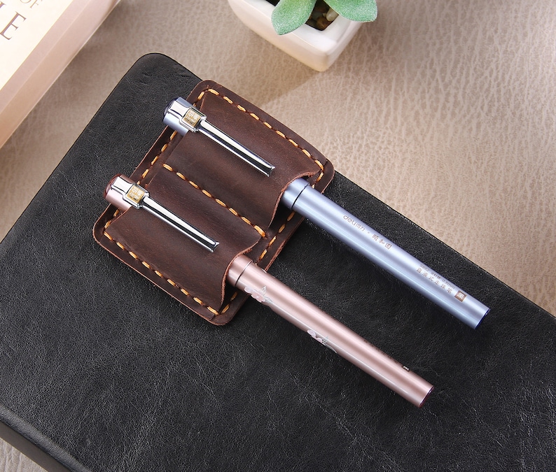 May include: A brown leather pen holder with two slots for pens. The holder has a stitched edge and is attached to a black notebook. Two pens are in the holder, one is silver and the other is rose gold.