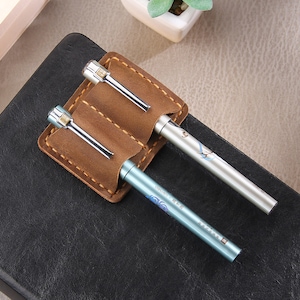 May include: A brown leather pen holder with two slots for pens. The holder is on a black surface and has two pens inside. One pen is silver with a blue and white design and the other is silver with a floral design.