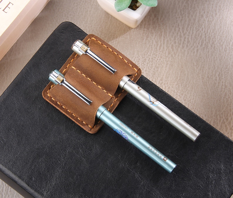 May include: A brown leather pen holder with two slots for pens. The holder is on a black surface and has two pens inside. One pen is silver with a blue and white design and the other is silver with a floral design.