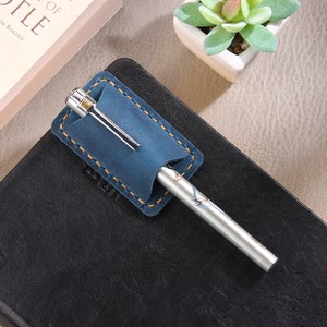 May include: A blue leather pen holder with a silver pen. The pen holder has a stitched edge and is attached to a black leather notebook.