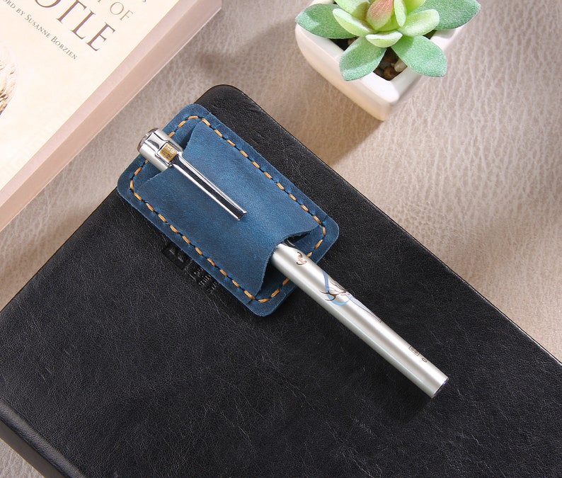 May include: A blue leather pen holder with a silver pen. The pen holder has a stitched edge and is attached to a black leather notebook.