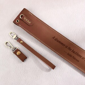 May include: Brown leather guitar strap with a quote that reads "A drummer is the backbone of any band." - Debbi Peterson. The strap has two brown leather keychains with gold metal rings.