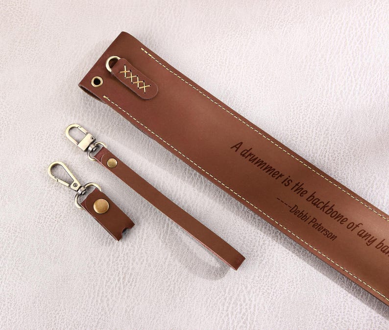 May include: Brown leather guitar strap with a quote that reads "A drummer is the backbone of any band." - Debbi Peterson. The strap has two brown leather keychains with gold metal rings.