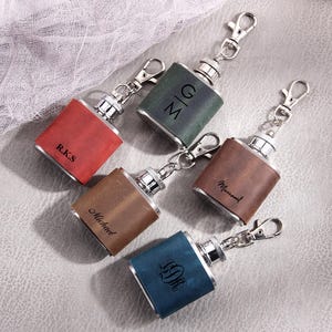 May include: Five leather-wrapped hip flasks with silver keychains. The flasks are personalised with the initials "R.K.S.", "Michael", "G|M", "Maxwell", and a monogram.