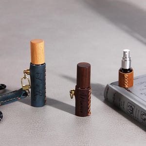 Peut inclure: Trois petits flacons pulvérisateurs cylindriques avec des bouchons en bois. L'un est gainé de cuir bleu avec le texte E & R, un autre de cuir marron foncé avec le texte Winnie, et un troisième de cuir beige. Chaque pièce présente des coutures.