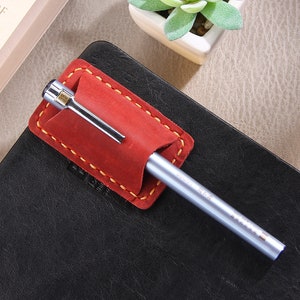 May include: A red leather pen holder with yellow stitching, attached to a black leather notebook. A silver pen is inserted into the holder.