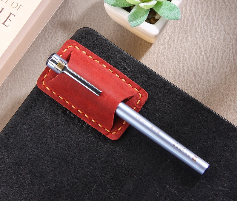 May include: A red leather pen holder with yellow stitching, attached to a black leather notebook. A silver pen is inserted into the holder.