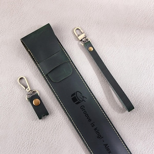May include: A dark green leather phone case with a gold metal keychain. The case has a drum symbol and the text "Groove is king! - Alex" embossed on it.