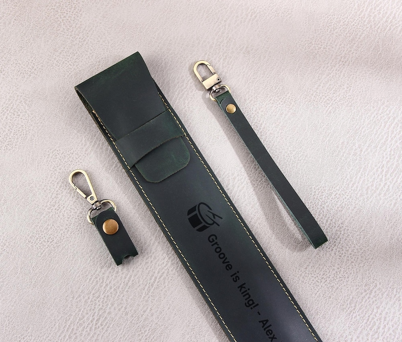 May include: A dark green leather phone case with a gold metal keychain. The case has a drum symbol and the text "Groove is king! - Alex" embossed on it.